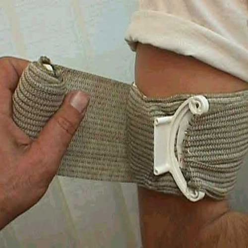 A person wraps a beige elastic bandage with a plastic fastener around their elbow for support. The bandage covers the joint area, and the persons thumb holds it in place while wrapping. The person is wearing a white shirt.