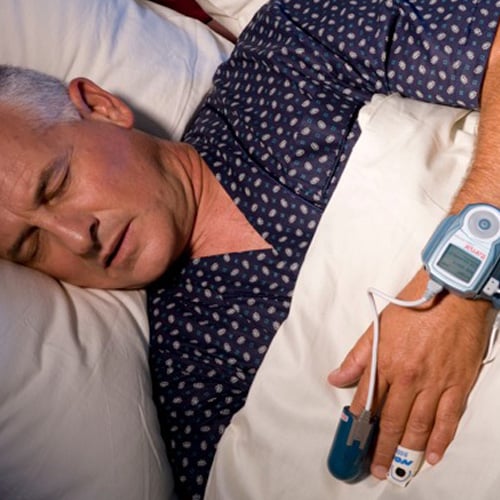 A closeup photograph of a man sleeping in bed with a Watch Pat devise on his left hand.