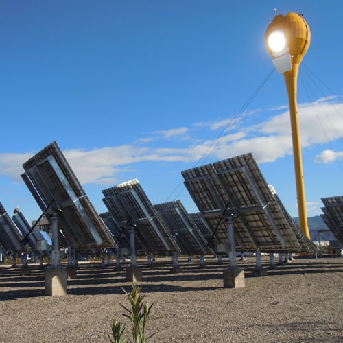 A group of solar panels sit in the middle of the desert ground, with a tall yellow sun collector that looks like the shape of a tulip in front of them. In the background, a clear blue sky emphasizes a sunny, clear day outside.