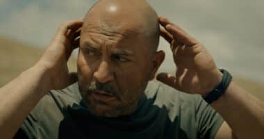 "Bald man with a beard outdoors under a cloudy sky, wearing a dark t-shirt and watch, holding his hands to his head and looking concerned or focused in a natural setting."