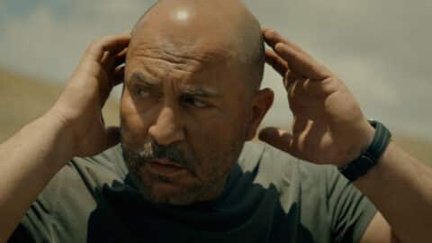 "Bald man with a beard outdoors under a cloudy sky, wearing a dark t-shirt and watch, holding his hands to his head and looking concerned or focused in a natural setting."