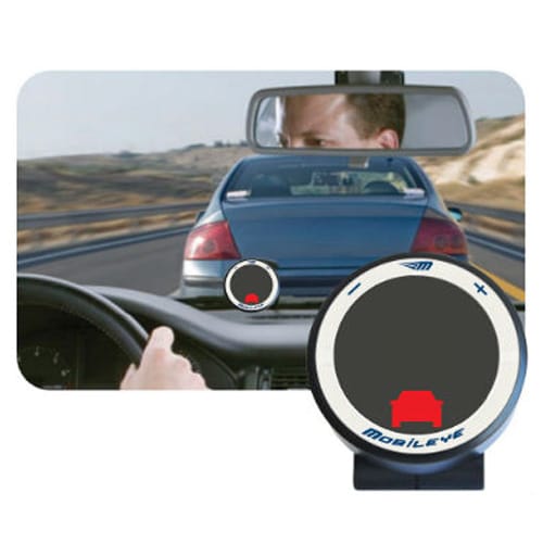 A car is driving closely behind another on a highway, as seen from the drivers perspective. The drivers hand is on the steering wheel. The rearview mirror shows part of the drivers face. A Mobileye device in the foreground displays a car icon.